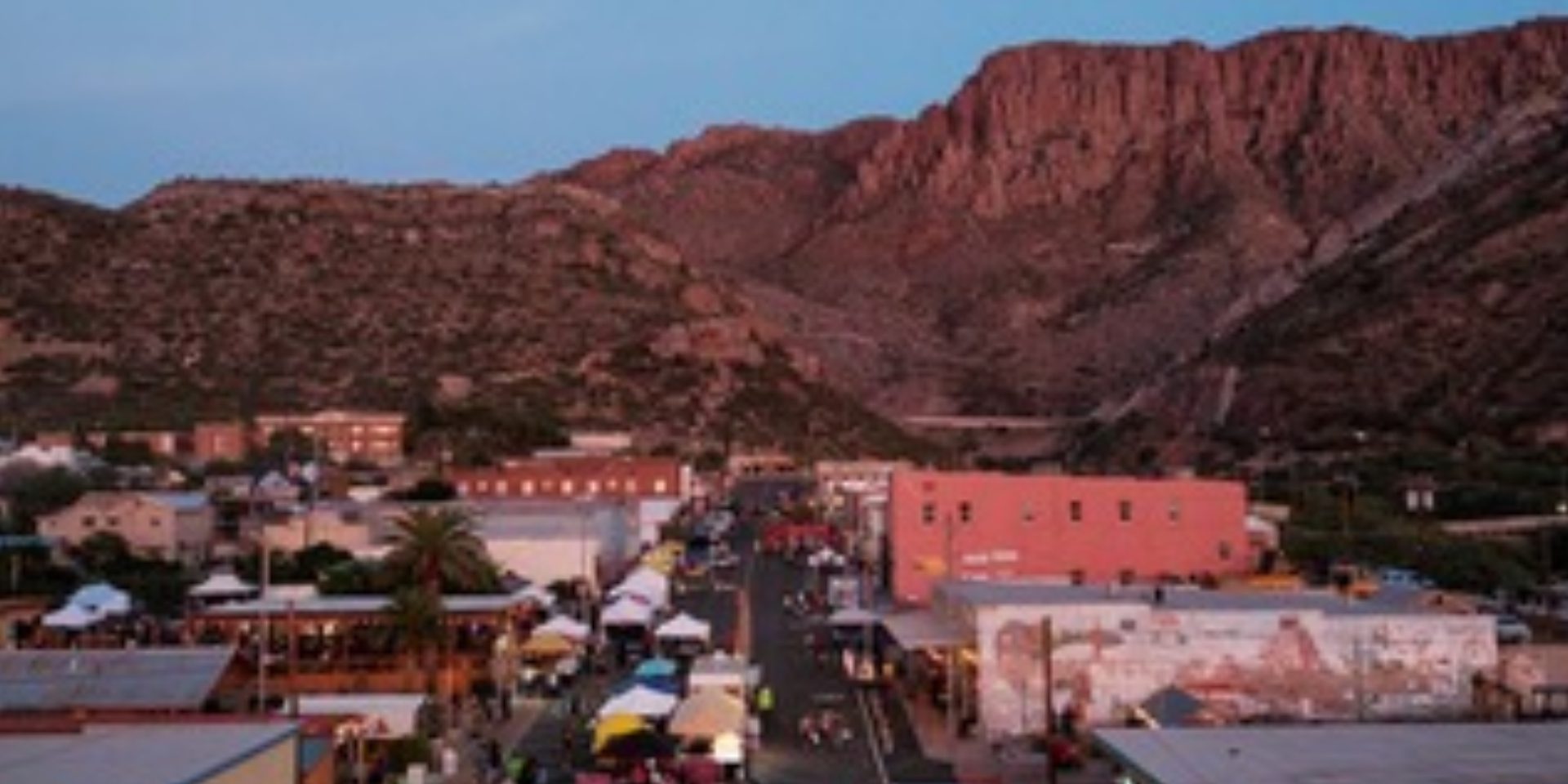 A street fair with tents and people is set up in a small town surrounded by rocky mountains at dusk.